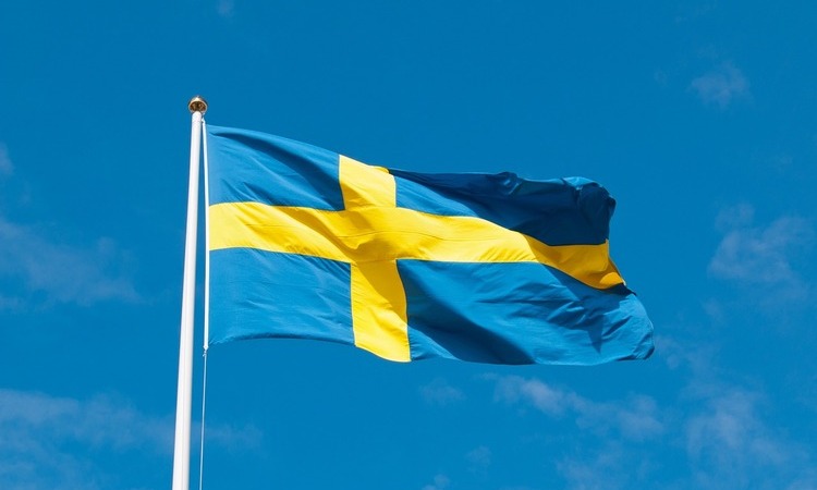 Sweden's national flag, blue with a yellow Nordic cross, waving against a clear blue sky.
