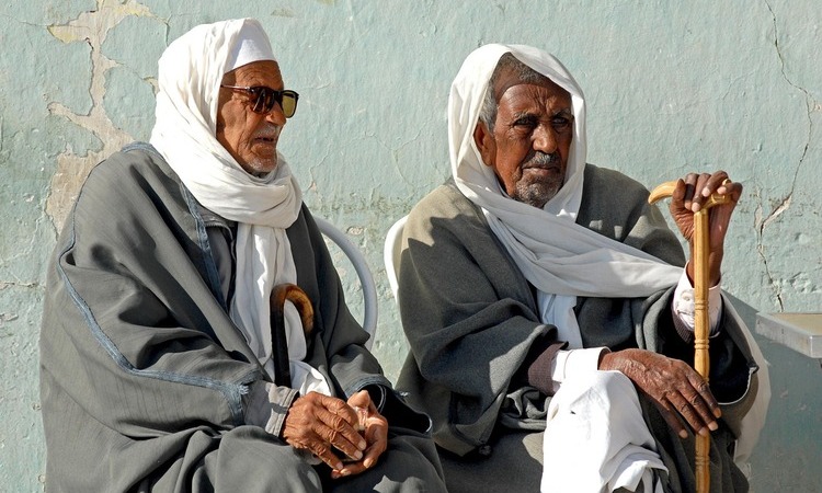 Two elderly Tunisian men wearing traditional robes and turbans sit outdoors.